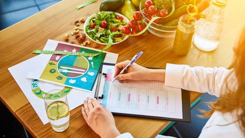 Woman dietitian in medical uniform with tape measure working on a diet plan sitting with different healthy food ingredients in the green office on background.