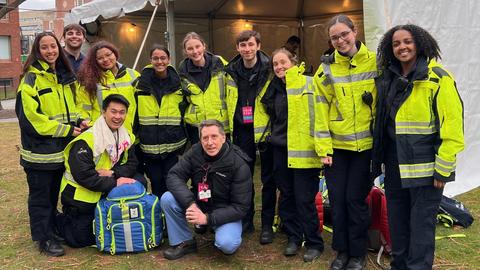 A diverse group of thirteen individuals in bright yellow emergency response jackets pose together in front of a white tent during an overcast day.