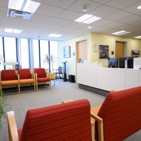 The Behavioral Health waiting and reception area with red and orange striped chairs, a bright yellow wall, a reception desk with computers, and large windows allowing natural light.