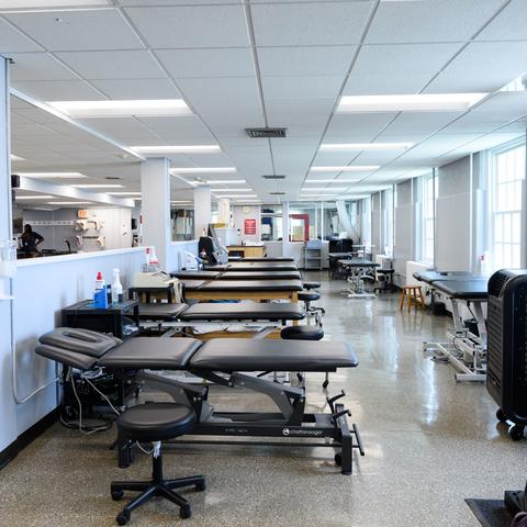 A bright physical therapy gym with multiple treatment tables, exercise equipment, and large windows, creating a clean and organized environment.