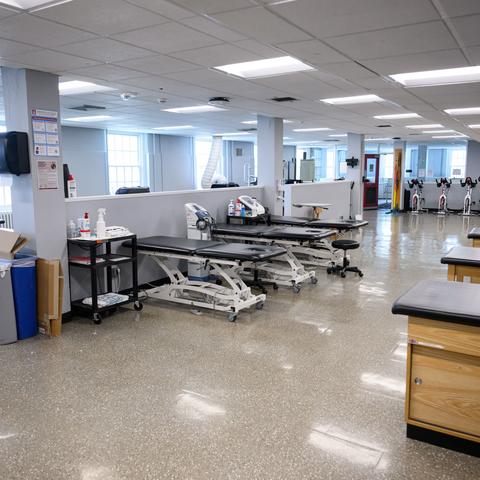 A spacious physical therapy room with treatment tables, a supply cart, and exercise bikes, illuminated by natural light through large windows.