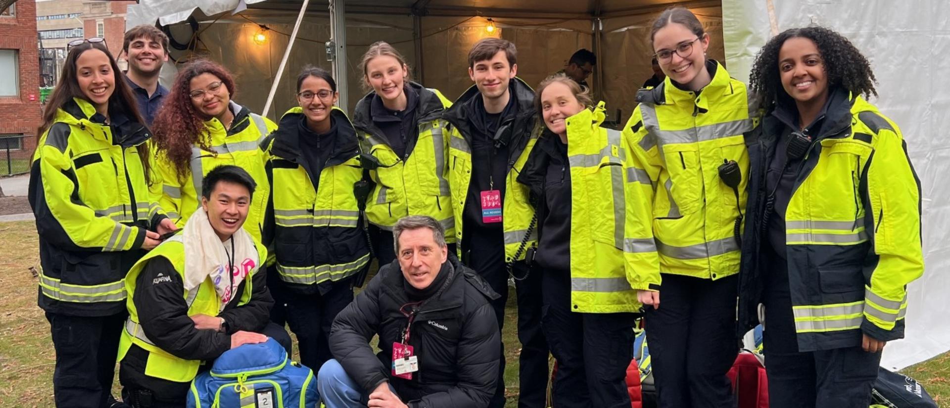 A diverse group of thirteen individuals in bright yellow emergency response jackets pose together in front of a white tent during an overcast day.