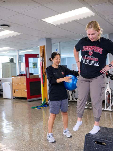 A trainer assists a woman balancing on one foot on a black exercise step in a gym, while she holds a blue exercise ball.