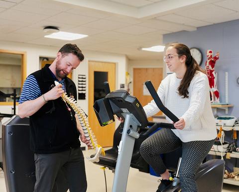 A man shows a spinal model to a woman on an exercise bike in the HUHS Physical Therapy area filled with medical equipment.