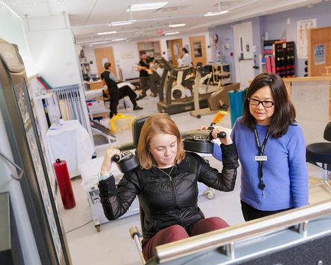 A young woman lifts weights on a machine in the HUHS physical therapy clinic while a staff member supports her and provides guidance. 